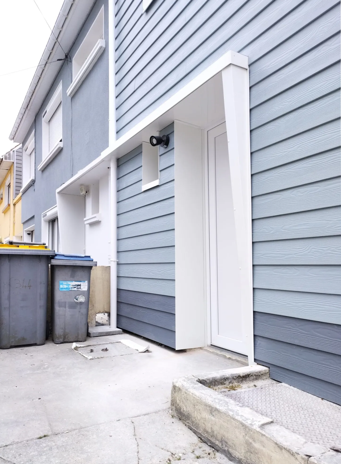 Entrée de la maison : bardage bleu-gris, porte blanche et applique extérieure